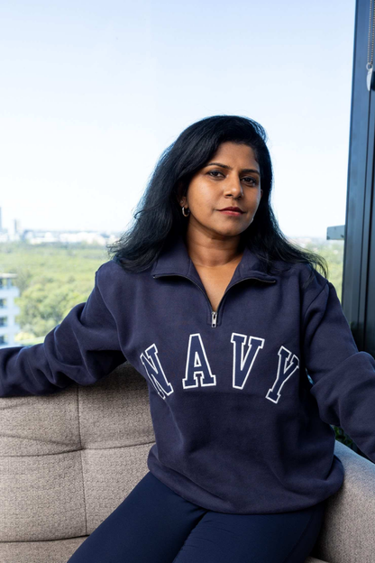 A woman wearing a navy blue quarter-zip sweatshirt with 'NAVY' printed in white varsity letters across the chest, sitting on a beige sofa in front of a large window.