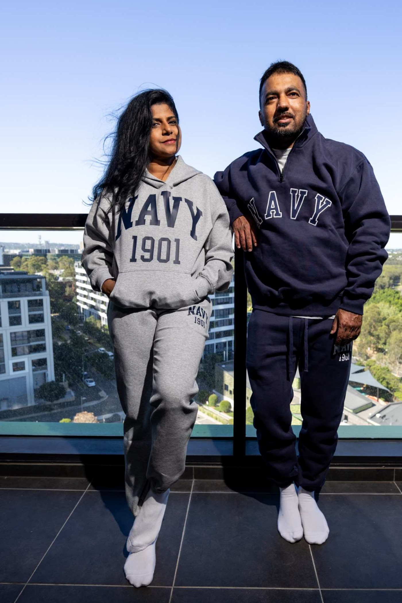 A man and a woman standing together on a balcony wearing matching navy and gray sweatpants and hoodies with 'NAVY 1901' printed on them, with a city view in the background.